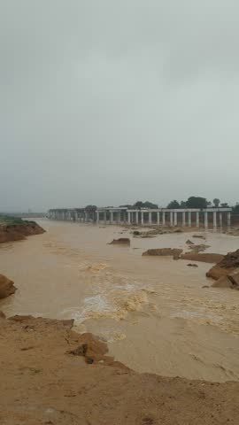 ​A wide shot of the Malir River in Karachi, Pakistan, during a flood. The brown, muddy water rushes forcefully past the damaged pillars of a collapsed bridge.