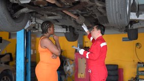 A professional senior Asian mechanic is explaining a vehicle problem by pointing to the car's undercarriage to a Black female customer in garage - Powered by Shutterstock - Get 15% off with code: PIKWIZARD15