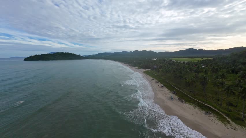 Low sunrise FPV aerial shot flying along the waves of Long Beach in San Vicente, Palawan. Calm tropical coastline with gentle surf, palm trees, and distant hills under morning cloud.