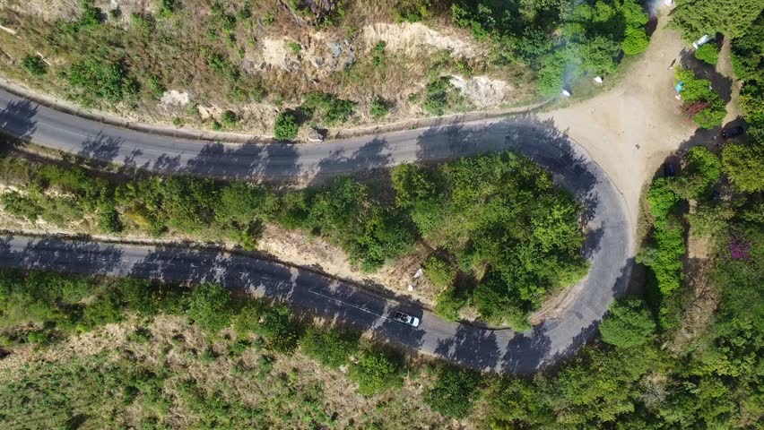 An aerial perspective reveals a winding road, a car, and lush vegetation under a sunny sky, with a rainbow adding a touch of magic to this picturesque scene.