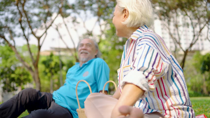 Senior asian man and woman couple of best friend enjoy a delightful picnic under the trees, sipping orange juice and sharing laughter in a park, creating lasting memories together, talk and relax