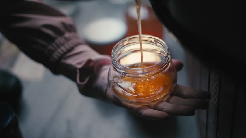 hand holding a glass jar, pouring and fill fresh golden extracted liquid organic honey into it, traditional honey production, apiculture and beekeeping in a bee farm 4k video