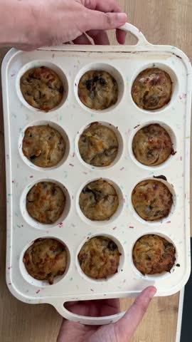 Close-up video of hands removing and showing freshly baked savory mushroom muffins with vegetables from a white silicone muffin tray on a wooden table, homemade food concept.