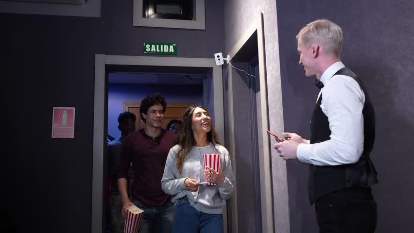 Multi ethnic groups of friends and an elderly woman entering a movie theater, receiving snacks and having their tickets checked by an usher, in slow motion