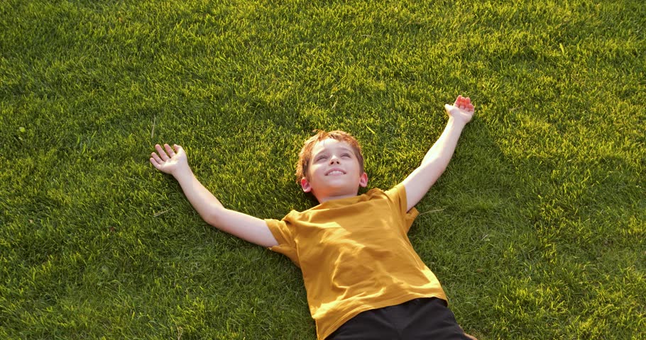 Happy child lies on green grass and looks at sky. child smiles and breathes deeply after play. Warm sunset light creates a peaceful joyful mood. childhood outdoors and joyful memories.