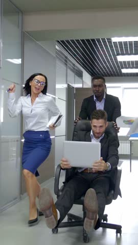 Group of professionals in a modern office setting, showcasing teamwork and collaboration, with one individual in a rolling chair using a laptop while others support and encourage the effort