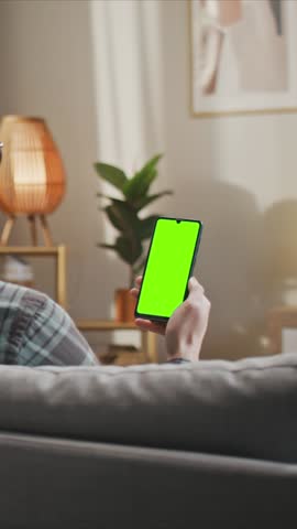 Close up male holding smartphone with chroma key green screen lying on sofa. Man using mobile device relaxing at home in living room. Vertical orientation.