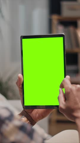 Man holding digital tablet with green screen close up, finger touching display, using chroma key mockup indoors at home. Male person interacting with blank screen device.