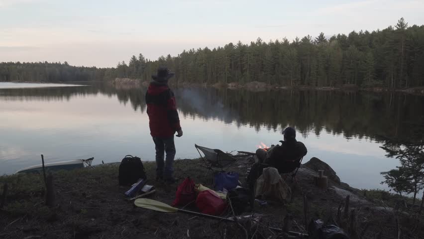 A peaceful lakeside scene with two Caucasian people relaxing by a campfire at sunset in the remote wilderness of Northern Ontario Crownlands