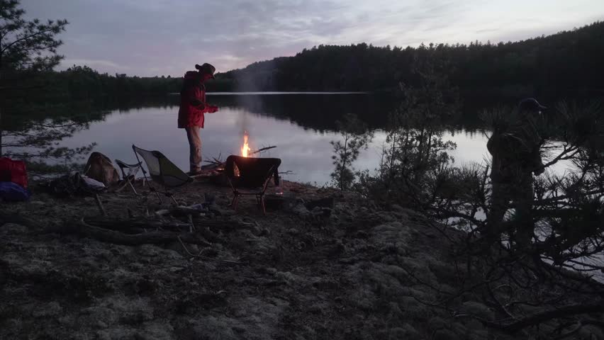 A quiet dusk scene in the Northern Ontario Crownlands tundra shows two Caucasian people by a campfire near a reflective lake surrounded by wilderness