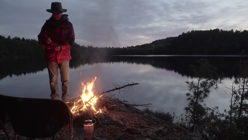 A man standing by a glowing campfire on the shore of a calm lake at dusk in the remote wilderness of Timmins, Northern Ontario, Canada