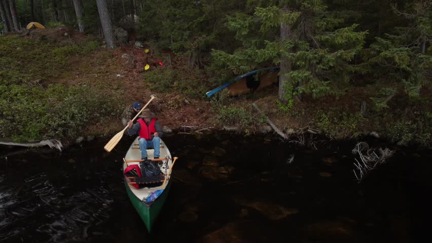 An aerial view of a man mooring a canoe at the shoreline of a dark lake in the Algoma Region of Northern Ontario Crownlands, Canada wilderness