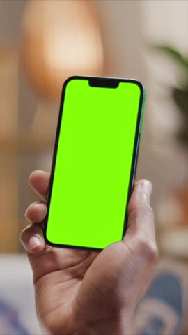 Close up African American male hand holding smartphone with chroma key green screen. Person showing mobile device mockup indoors for browsing, apps, social media use. Vertical format.