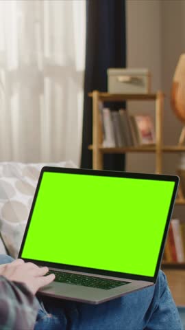 Man sitting with laptop on knees, green screen display for replacement, working or browsing at home on bed. Male person using computer indoors, mockup screen for app or website design.