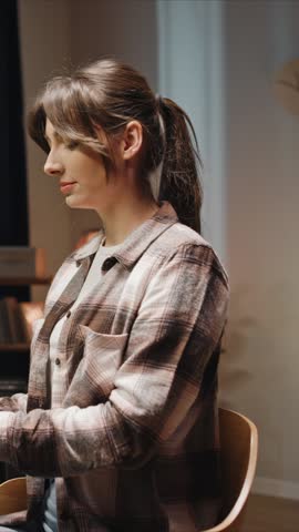 Side view of woman sitting at desk indoors, wearing plaid shirt, using computer. Portrait of happy smiling female concentrating on gadget while working, browsing, or studying at home.
