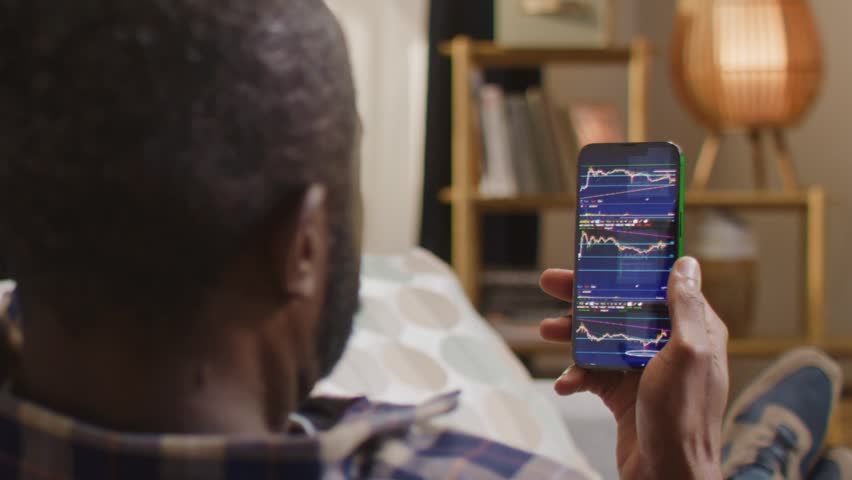 African American man holding smartphone with stock market app open, financial charts and data on screen, sitting at home on couch. Male person tracking investments using mobile trading application.