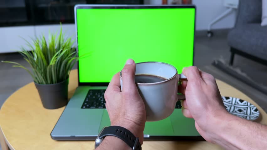 4K video of hands holding a coffee mug in front of a green screen laptop. Ideal for lifestyle edits, productivity mockups, or chroma key compositing in cozy work-from-home scenes.