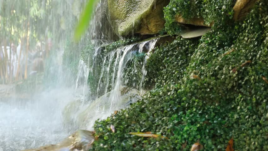 Close-up of decorative waterfall and lush foliage in a botanical garden, with detailed view of water streams, mossy stones and ornamental design elements