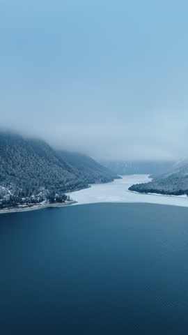Flying over the wide grey waterscape of the lake surrounded by mighty mountains. Tops of the rocks are coated in grey cloudscape. Aerial view. Vertical video.