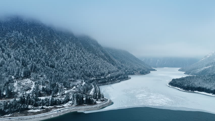 Snow-sprinkled pine woods growing on the slopes of the gorgeous mountains. Drone footage over the lake covered with white ice.