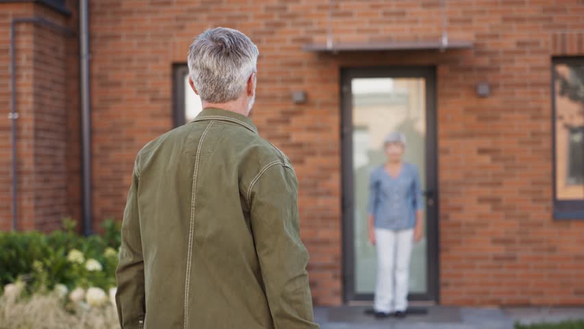 Man with gray hair spreading arms wide standing on path in front of brick house. Woman walking toward him with open hands ready for hug. Couple enjoying reunion moment together outdoors.