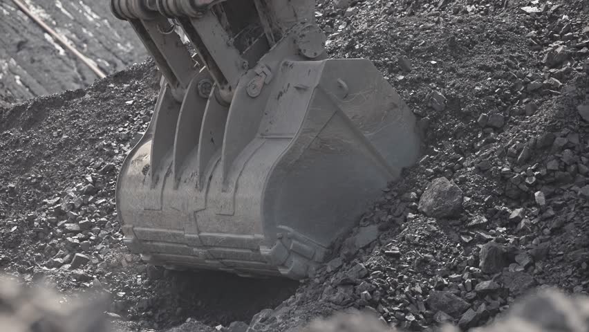 Bucket of mining excavator scoops up coal from mine deposit and loading into back of yellow dump truck.