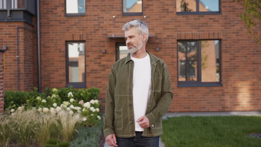 Smiling mature man with gray beard standing arms crossed in front of brick building. Male looking at camera with relaxed confident pose. Outdoor portrait showing happiness near modern house.