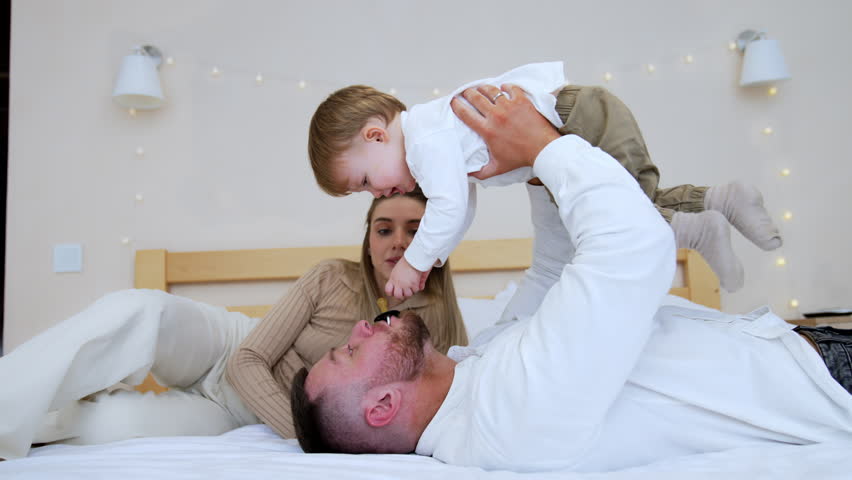 Bearded man lying on the bed holds his son above. Dad plays with a kid. Woman lying behind looks at her loved ones.