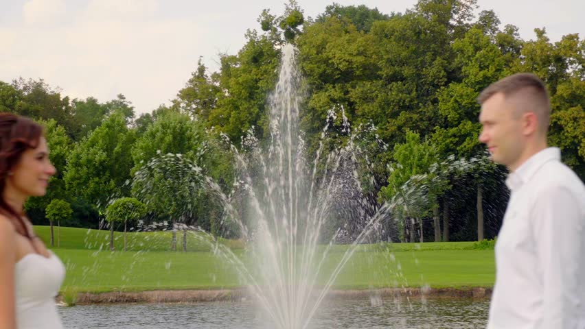 Romantic Young Adult Couple Groom and bride embrace exchanging a kiss with a park fountain and greenery as the scenic backdrop, symbolizing love and passion in a picturesque outdoor setting 4K UHD