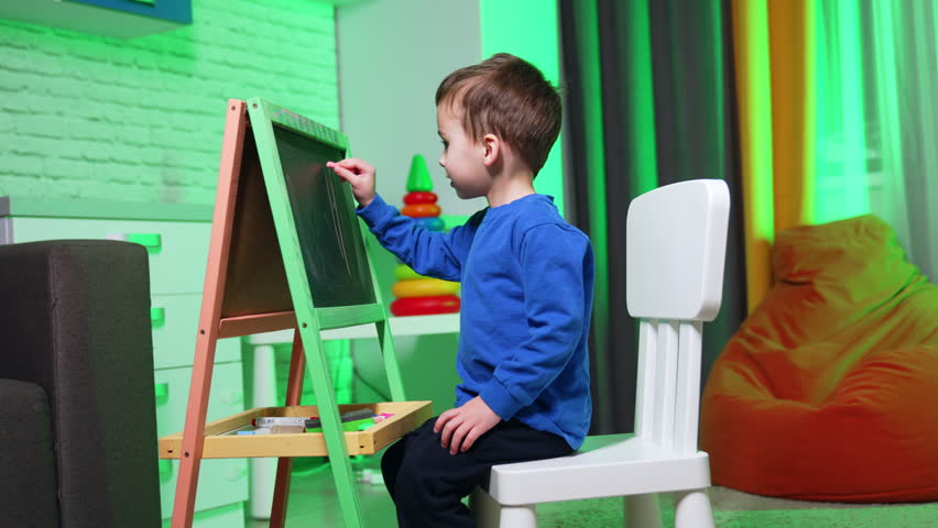 Lovely baby boy sits at the blackboard drawing with chalk. Cheerful kid having fun at home.