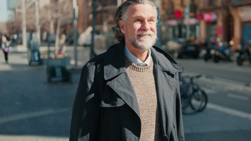 Smiling senior man walking down a city street on a sunny day. He is wearing a stylish coat and sweater and has long gray hair and a beard.