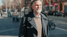 Smiling senior man walking down a city street on a sunny day. He is wearing a stylish coat and sweater and has long gray hair and a beard. - Powered by Shutterstock - Get 15% off with code: PIKWIZARD15