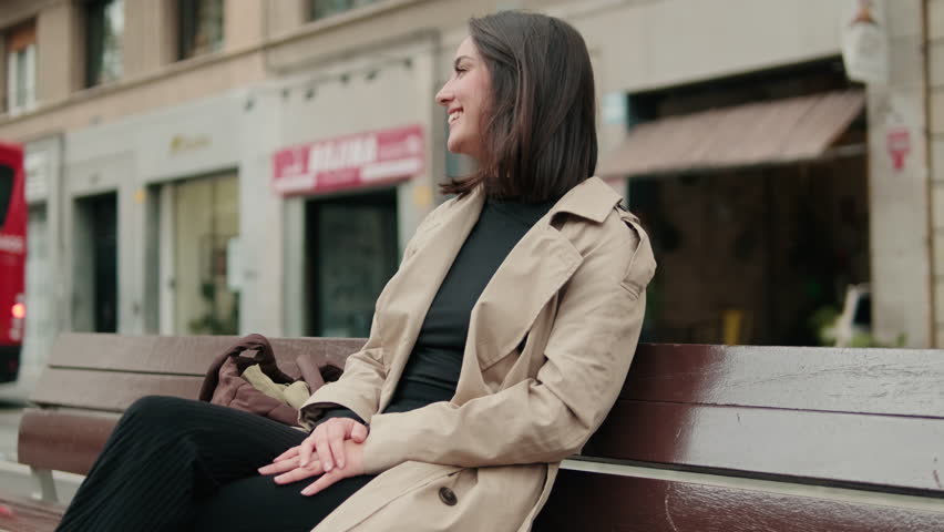 Young woman sits on a bench in the city, smiles and looks around. She