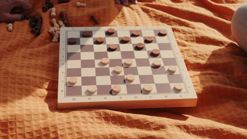 Friends playing checkers on a picnic blanket outdoors. Sunlight creates long shadows over the blanket and game.