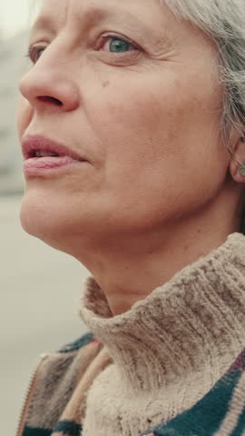 Vertical video, A mature woman with grey hair and blue eyes, stands on a city street corner. She wears a brown turtleneck sweater, plaid jacket, and small pearl earrings.