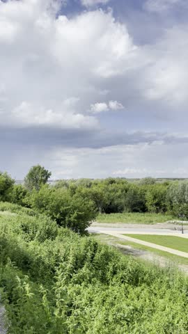 Cinematic aerial view of urban riverside promenade with lush greenery, vintage street lamps, geometric landscaping, dramatic cloudy sky, vibrant summer colors, riverside atmosphere for Reels, Stories.