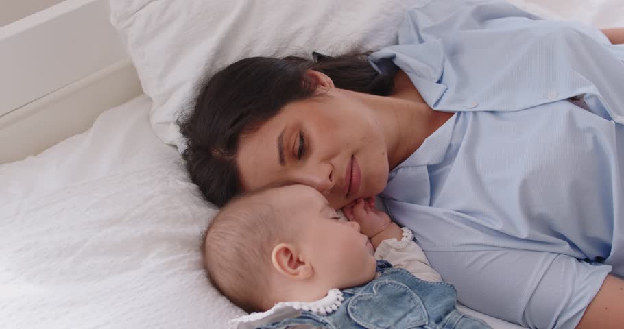 Portrait of smiling young happy mother lying on bed near cute little newborn child and admiring her sleeping baby in bedroom at home. Family, love, care and mothers day concept. 4k video.