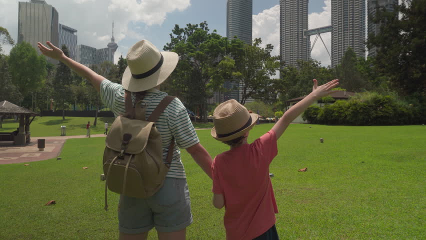 Female tourist and her son are raising their arms and enjoying cityscape in Kuala Lumpur, Malaysia. Woman and little boy in hats at a city park among skyscrapers. Freedom concept. Traveling with child