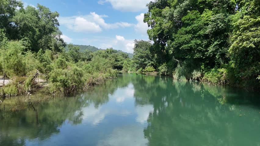 Low-altitude drone flight upstream on the Tenexio River in Jalpilla, Axtla de Terrazas, Huasteca Potosina, Mexico, with flowing water, blue sky, white clouds, and lush green scenery.
