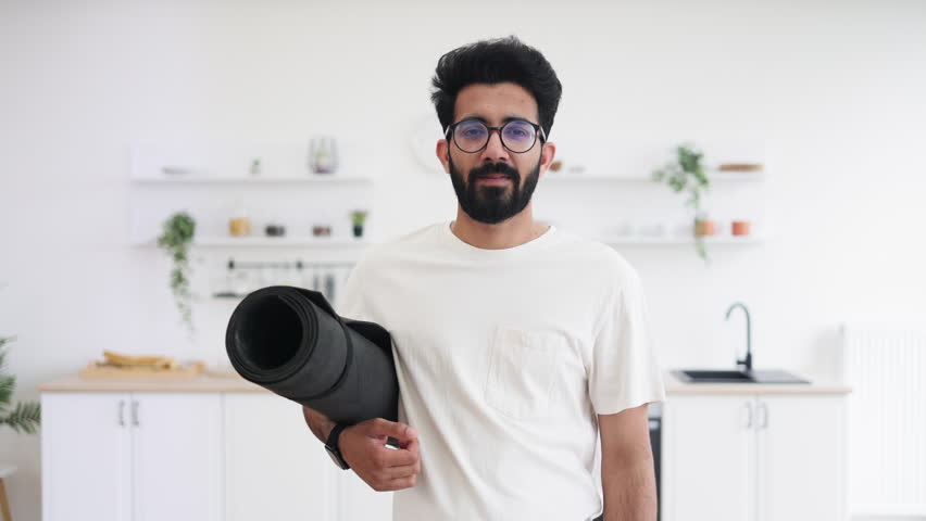 Young adult man, South Asian appearance, smiling, standing in kitchen holding yoga mat, fitness context