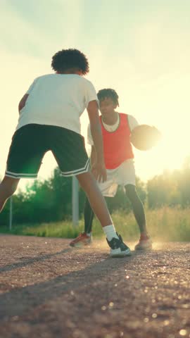 Two teenagers playing basketball on outdoor court in summer, vertical view. Sportive hobby for young people, adolescents practicing streetball, cinematic slow motion shot, do sport and be active
