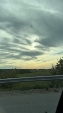 Vertical road trip video with cloudy dramatic sky at sunset, trees and roadside guardrail filmed from moving car, travel and adventure mood.