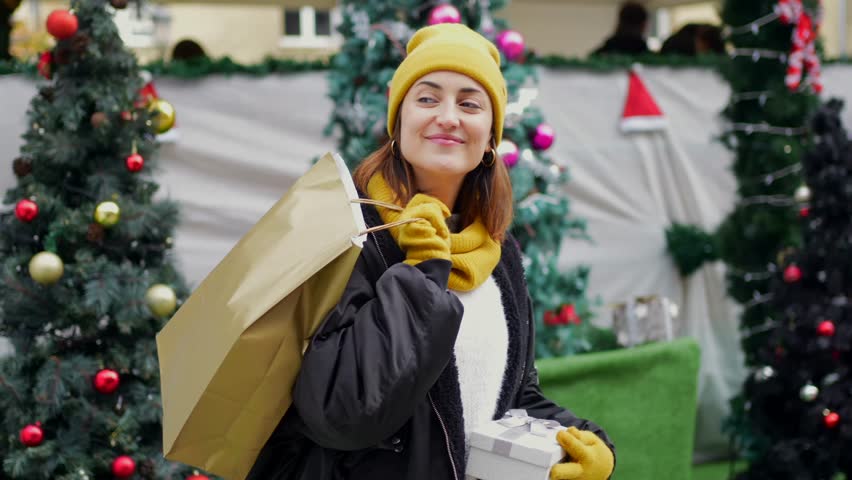 Cheerful young woman wearing winter attire, holding shopping bag and wrapped gift while standing near decorated christmas market with festive lights and tree