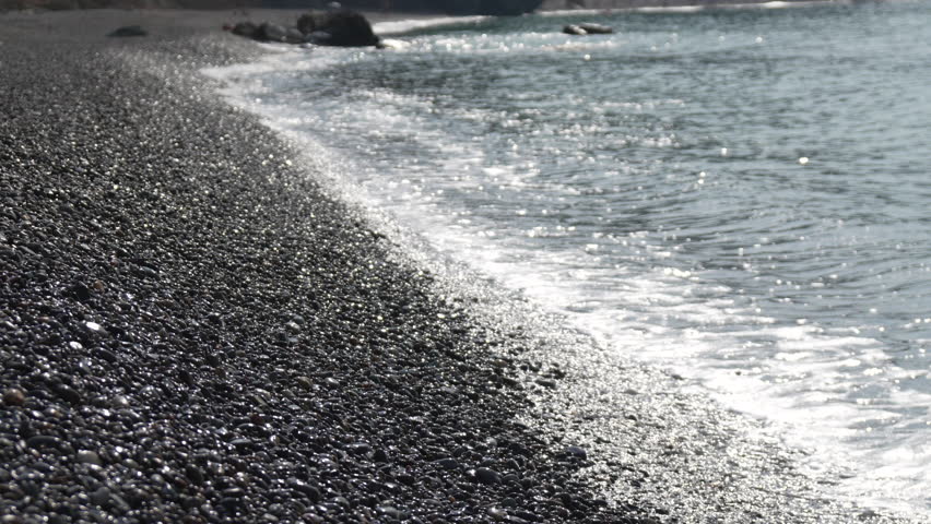 Pebble beach, ocean waves, sparkling water gently washing black stones on a sunny day.