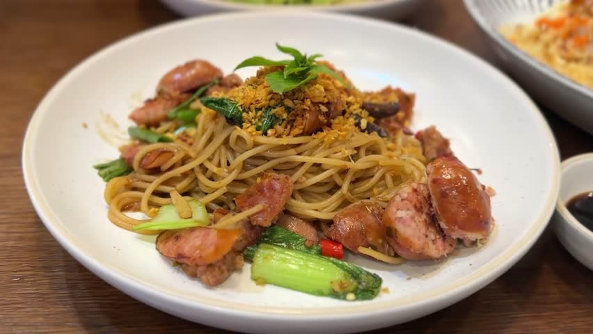 Close-up of spicy spaghetti with duo sausages, crispy garlic, and vegetables on white plate.