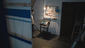 Young man operates touchscreen control panel in low-light studio near wall with messy notes, diagrams, and plugged-in cords while surrounded by technical equipment - Powered by Shutterstock - Get 15% off with code: PIKWIZARD15