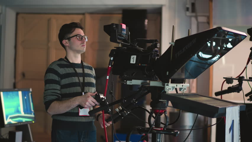 Man operating professional TV camera with teleprompter in broadcast studio. Monitor shows studio backdrop. Operator wearing striped sweater and glasses, engaged in filming process