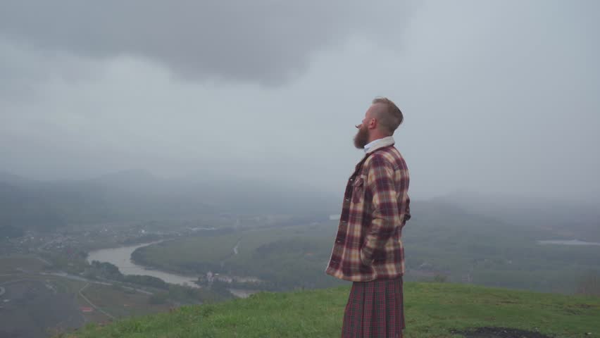 An adult redhead bearded man in Scottish national dress plaid jacket and kilt stands on a hill and looks into the distance. Masculinity, pride and human dignity concept.