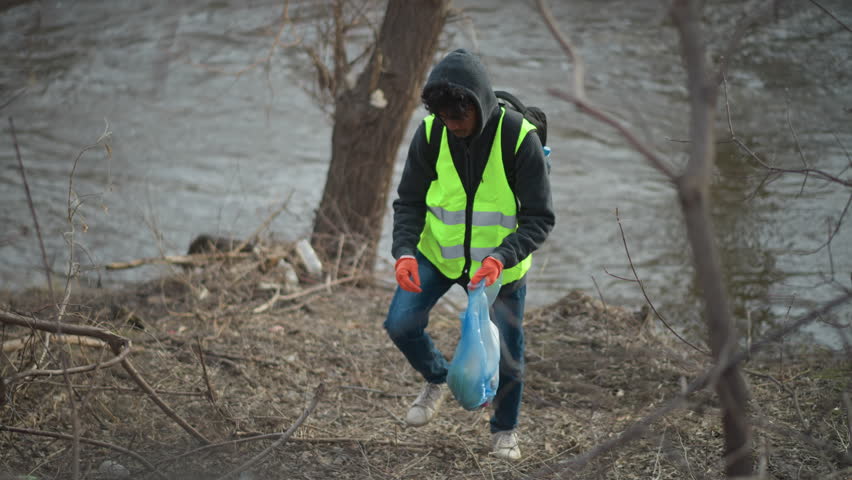 Volunteer in reflective vest and hoodie with backpack and gloves picks up litter along riverbank, placing trash into blue plastic bag during environmental cleanup to protect nature