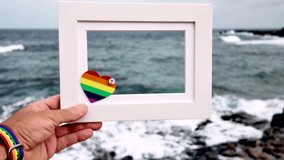 Slow motion video of hands holding a white frame with a small rainbow heart, celebrating gay pride and love with the ocean waves in the background in the Canary Islands - Powered by Shutterstock - Get 15% off with code: PIKWIZARD15